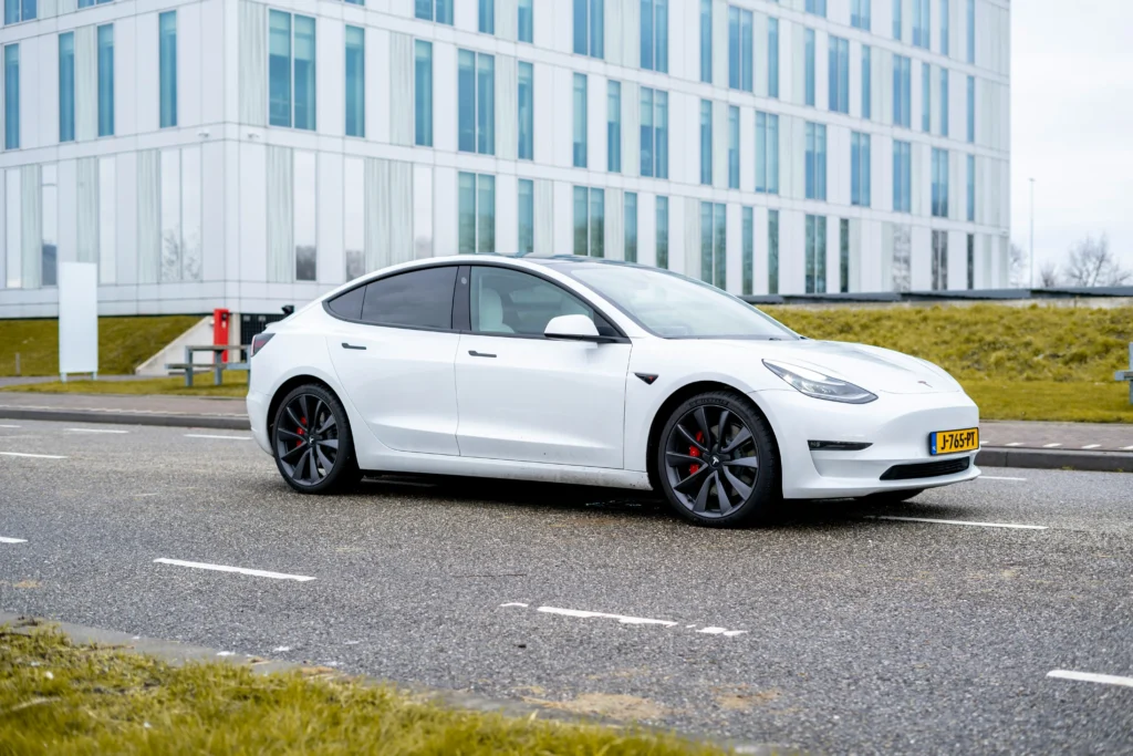 White Tesla Model 3 electric car on asphalt road in front of modern building.